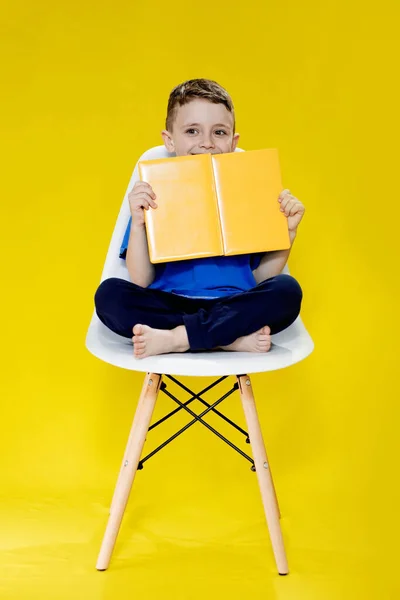少し陽気なブロンドの緑目の男の子5 6歳でスタイリッシュな青のTシャツを保持本と黄色の壁の背景に読書 子供のスタジオの肖像画 — ストック写真