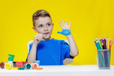 Küçük bir anaokulu öğrencisi elinde fırçayla masada oturur ve avucunu boyar. Anaokulu geliştirme