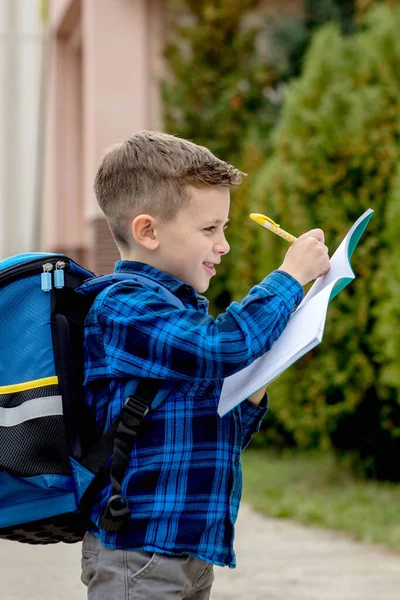 学校の庭でノートにメモを書いて笑顔で バックパックとカジュアルな服を着た幸せな少年 — ストック写真