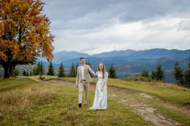 A married couple walks among the mountains, the bride leads the groom behind her, the girl holds the boy's hand, smiles, the woman in a white wedding dress, the groom and a beige suit.