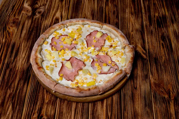 Delicious pizza with ham, pineapple, corn on a creamy base on woden background. Hawaiian pizza.