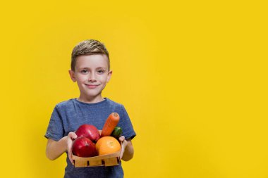 Çocuk sarı arka planda taze sebzeler ve meyvelerle dolu bir sepet tutuyor. Vegan ve sağlıklı konsept.
