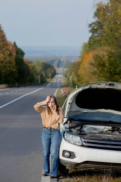 若い女性ドライバーを動揺彼女の車に問題があるエンジンを検査しながら オープンフードを持つ壊れた車の近くの支援サービスと携帯電話で怒って話す — ストック写真