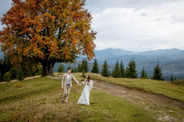 A married couple walks among the mountains, the bride leads the groom behind her, the girl holds the boys hand, smiles, the woman in a white wedding dress, the groom and a beige suit.