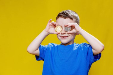 Mavi tişörtlü küçük mutlu anaokulu çocuğu sarı arka planda gözlerinin önünde ethereum ve bitcoin paralar tutuyor..