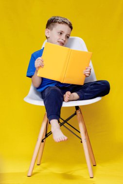 Küçük neşeli, yeşil gözlü çocuk. 5-6 yaşlarında, şık mavi tişörtlü, elinde kitap olan ve sarı duvar arkasında okuyan çocuk stüdyosu portresi..