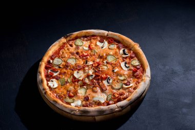 Appetizing pizza with hunting sausages, mozzarella cheese, pickled cucumbers, chili peppers on a tomato base on black background.