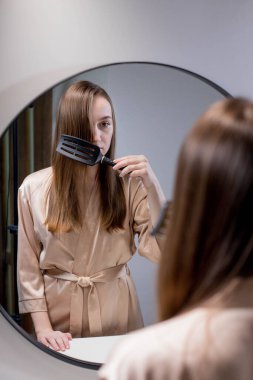 Güzel bir kadın, güzel saçlarını fırçayla tararken banyoda aynanın yanında duran çekici genç bir bayan yansımasına ve gülüşüne bakarak boş alanla seçici bir şekilde odaklanıyor. Saç bakımı konsepti.