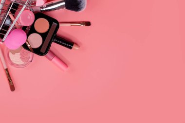 Pembe arka planda makyajlı alışveriş arabasıyla yaratıcı bir konsept. Parfüm, sünger, fırça, rimel, göz farı, sepetteki dudak parlatıcısı..