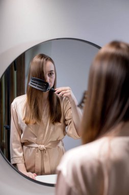 Güzel bir kadın, güzel saçlarını fırçayla tararken banyoda aynanın yanında duran çekici genç bir bayan yansımasına ve gülüşüne bakarak boş alanla seçici bir şekilde odaklanıyor. Saç bakımı konsepti.