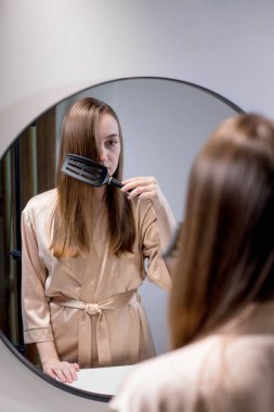 Güzel bir kadın, güzel saçlarını fırçayla tararken banyoda aynanın yanında duran çekici genç bir bayan yansımasına ve gülüşüne bakarak boş alanla seçici bir şekilde odaklanıyor. Saç bakımı konsepti.