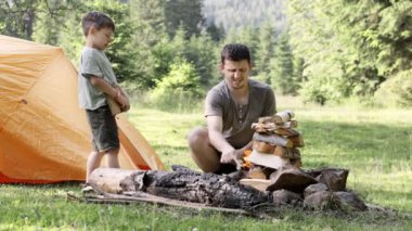 Baba ve oğlu yakacak odun yığınının yanında yan yana oturur ve çadırın yanında ateş yakarlar. Macera ve macera ruhu. Yürüyüş ve kamp. Seyahat konsepti 4K.
