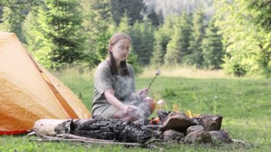 Bir kadın ormanda şenlik ateşinde şekerleme pişiriyor. Sıcak bir tatlı çubuğuna üfle. Doğada piknik yaparız..