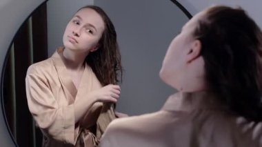 Genç bir kadın banyoda aynanın önünde dikilirken saçlarını havluyla siler..