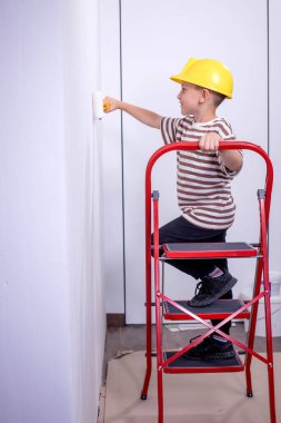 Bir çocuk müteahhit merdivenin üzerinde durur ve bir apartmanın beyaz duvarlarını boyar. Tadilat sırasında boya silindirli bir çocuk..