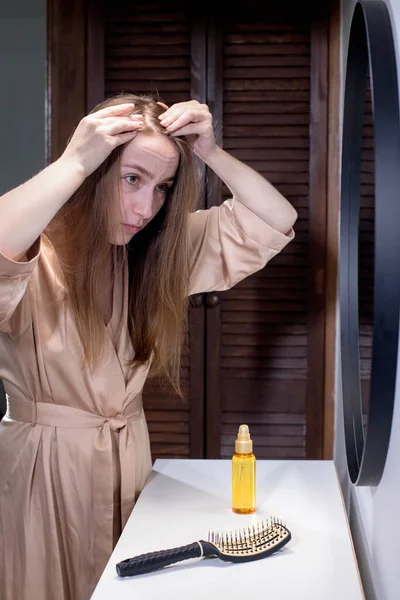 Hair loss. Depressed young woman looking at her hairbrush and expressing negativity while standing against mirror.