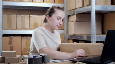 İnternetteki kadın depo işçisi dizüstü bilgisayar kullanıyor. Paket kutuları hazırlıyor. Çevrimiçi perakende satış işlemlerini kontrol ediyor.