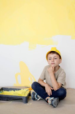 İnşaat üniformalı bir çocuk boyaların yanında oturuyor. Onarım. Duvarları boyuyorum..