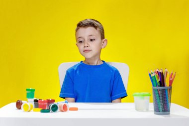 Bir öğrenci boyalara bakar ve hangi rengi seçeceğini düşünür. Çocuk elinde boya ve kalemlerle masada oturuyor.