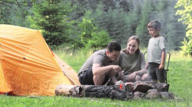 Piknikte çocuklu mutlu bir aile çadırın yanındaki ateşin yanında oturur ve ormanda kamp ateşinde yemek pişirir. Kamp, dinlenme, yürüyüş..