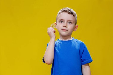 Little happy preschool boy in a blue t-shirt holding bitcoin in front of his eyes on yellow background.