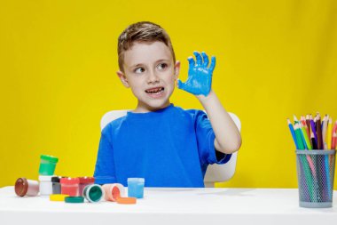 Küçük bir anaokulu öğrencisi masada oturuyor, avuç içlerini bir kağıt parçasına çiziyor. Renkli el izleri. Anaokulu geliştirme.