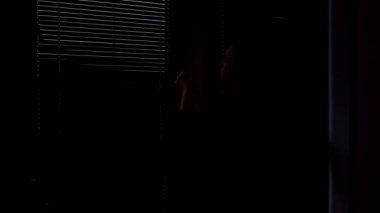 Perdeleri açan bir kadının silüeti. İç taraftaki büyük pencerelerde açılan tahta panjurlar..