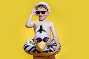 Yaz panamalı çocuk ve sarı arka planda şişirilebilir yüzme çemberi olan güneş gözlüklü..