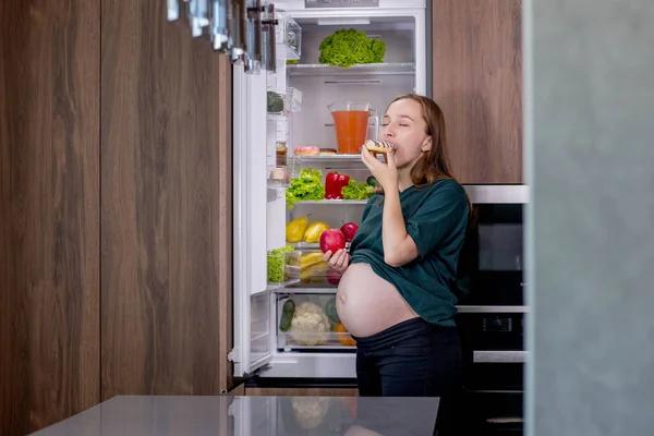 Pregnant woman near the refrigerator eating dessert.