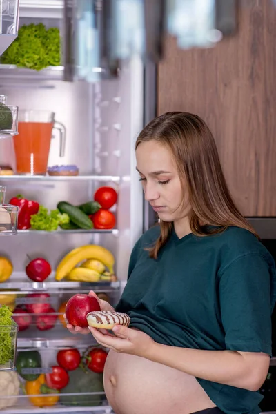 Pregnant woman near the refrigerator chooses what to eat between vegetables and dessert.