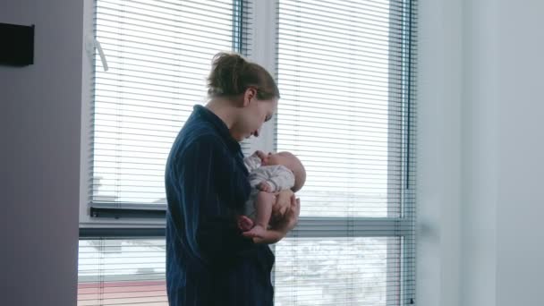 美しい若い女性は 自宅の窓の隣に立っている間 彼女の腕に赤ん坊を抱いています かわいい リトル トッドラー — ストック動画