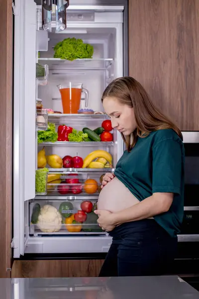 Hungry pregnant woman standing near refrigerator looking for food during pregnancy. Healthy eating.