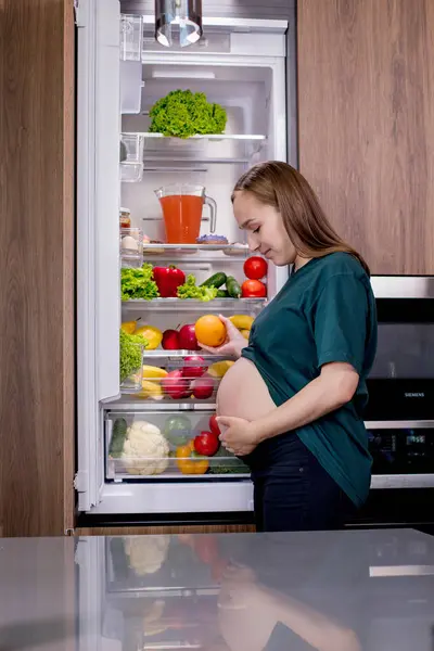 Young pregnant woman with an orange near the refrigerator at home. Healthy Eating.