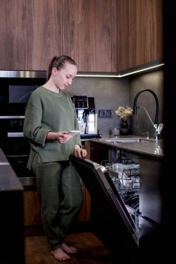 Kadın elinde akıllı ev programı çalışan bir telefon tutuyor, bulaşık makinesini uzaktan kontrol ediyor. Evde akıllı mutfak kavramı..