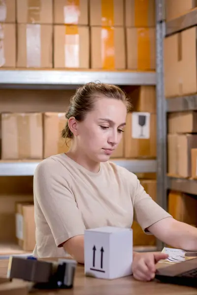 Portrait of a businesswoman holding a box and typing something on a keyboard while preparing package information. Postal service and small business concept.