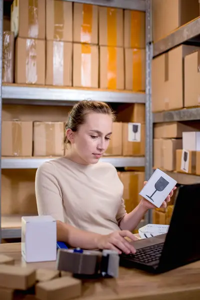 Portrait of a businesswoman holding a box and typing something on a keyboard while preparing package information. Postal service and small business concept.