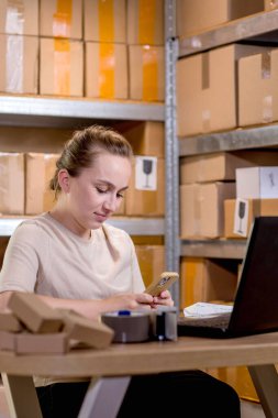 Evde çalışan genç girişimci küçük işletme akıllı telefondan internet siparişlerini kontrol ediyor ve müşterilere teslim edilecek kutulu eşyalar hazırlıyor.