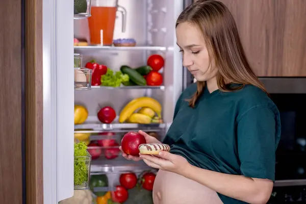 冷蔵庫の近くの妊婦は 野菜とデザートの間で何を食べるかを選択する ロイヤリティフリーのストック写真