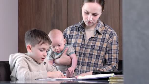 Mutter Mit Baby Hilft Ihrem Ältesten Sohn Bei Den Hausaufgaben — Stockvideo