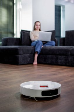 Genç bir kadın dizüstü bilgisayarda çalışıyor, koltukta oturuyor ve evdeki elektrikli süpürgeyi temizliyor. Akıllı ev aletleri ve evden çalışma kavramı.