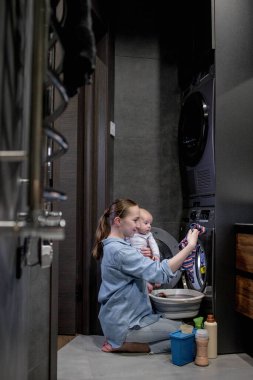 Çocuk kıyafetlerini yıkıyorum. Bir kadın, kollarında bir çocukla ev işi yapar..