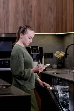 Kadın elinde akıllı ev programı çalışan bir telefon tutuyor, bulaşık makinesini uzaktan kontrol ediyor. Evde akıllı mutfak kavramı..