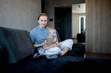 Anne karanlık bir koltukta oturup çocuğunu tutuyor ve evde astım spreyini kullanmalarına yardım ediyor.