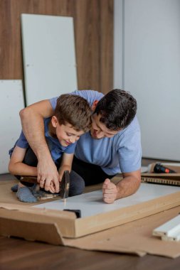 Oğlu, babasının mobilya toplamasına yardım ediyor. Yakınlarda da alet ve tahtalar var. Evde mobilya montajı.