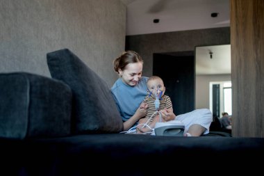 Anne karanlık bir koltukta oturup çocuğunu tutuyor ve evde astım spreyini kullanmalarına yardım ediyor.