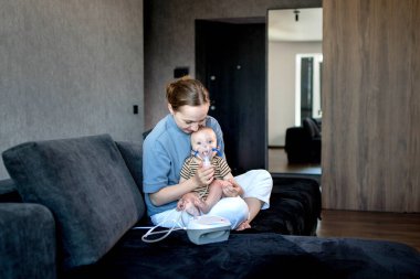 Anne karanlık bir koltukta oturup çocuğunu tutuyor ve evde astım spreyini kullanmalarına yardım ediyor.