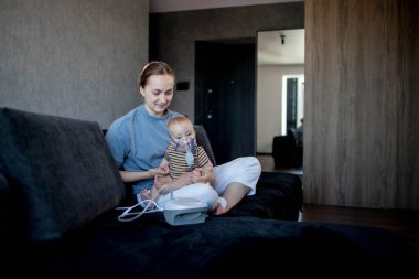 Anne karanlık bir koltukta oturup çocuğunu tutuyor ve evde astım spreyini kullanmalarına yardım ediyor.