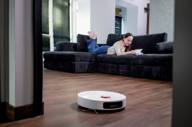 Woman lying on sofa and reading a book while robot vacuum cleaner doing housework. clipart