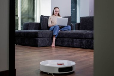 Genç bir kadın dizüstü bilgisayarda çalışıyor, koltukta oturuyor ve evdeki elektrikli süpürgeyi temizliyor. Akıllı ev aletleri ve evden çalışma kavramı.
