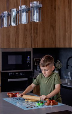 Küçük çocuk masaya merdaneyle zencefilli ekmek hamuru yuvarlıyor.